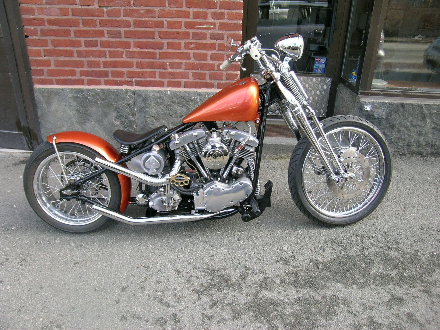 An orange motorcycle with a custom round oil tank featuring a spade and the number 13 cut into the metal.